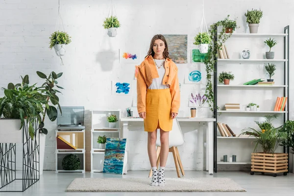 Hermosa mujer joven de pie en una amplia habitación decorada con plantas verdes y pinturas en la pared blanca - foto de stock