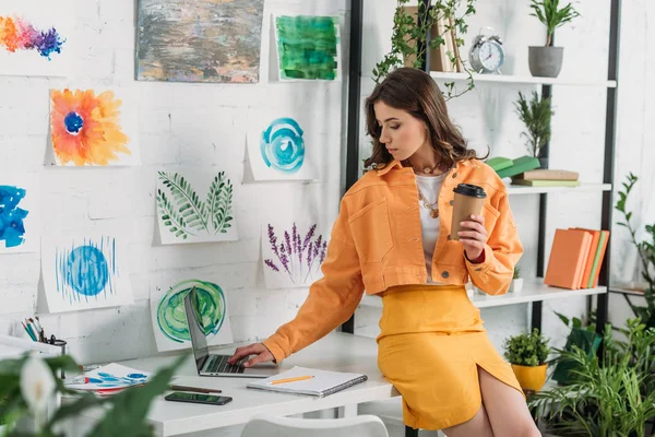 Pretty girl using laptop and holding paper cup while sitting on desk in room decorated with green plants and paintings on wall — Stock Photo