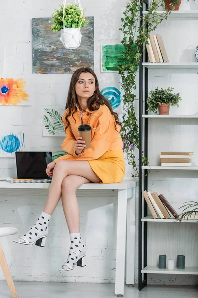 Chère fille assise sur le bureau près d'un ordinateur portable avec écran vierge, tenant la tasse jetable et regardant loin — Photo de stock