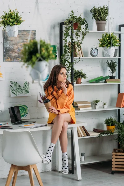 Pensive young woman sitting on desk in room decorated with green plants and paintings on wall — Stock Photo