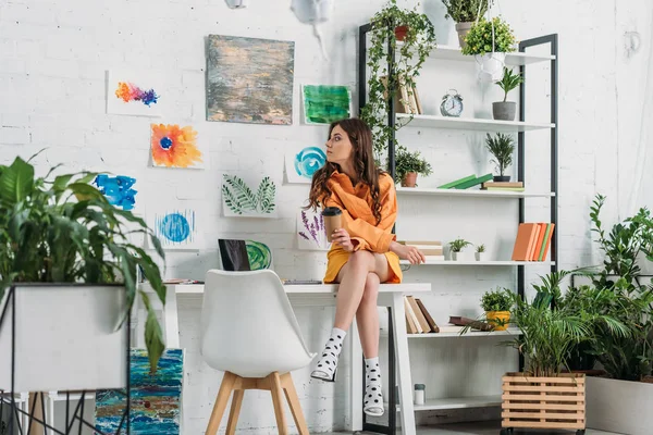 Jolie fille assise sur le bureau et tenant tasse en papier dans la chambre décorée avec des plantes vertes et des peintures sur le mur — Photo de stock