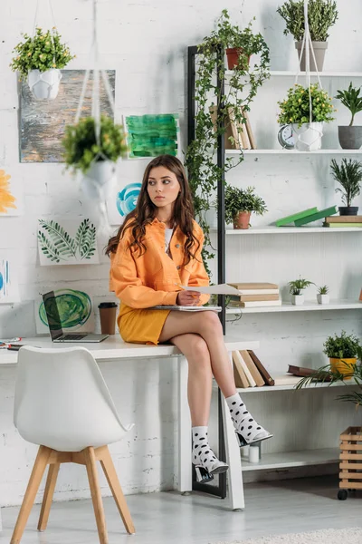 Задумчивая молодая женщина сидит на столе в комнате, украшенной зелеными растениями и картинами на стене — стоковое фото