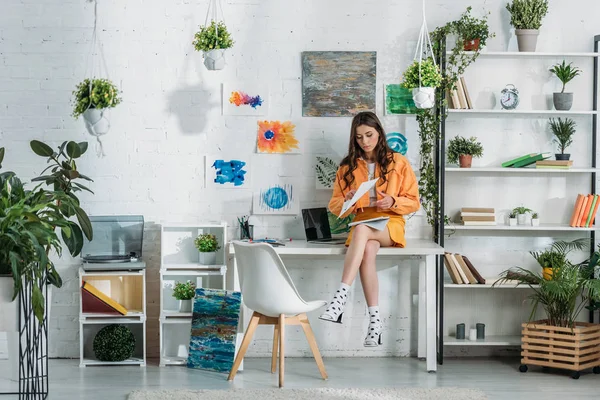 Thoughtful young woman sitting on desk in light spacious room decorated with green plants and paintings on wall — Stock Photo