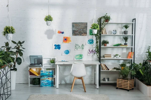 Luz quarto espaçoso decorado com plantas e pinturas na parede branca com prateleiras, mesa e cadeira — Fotografia de Stock