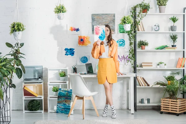 Trendy young woman covering face with painting while standing in spacious room decorated with green plants and drawings on white wall — Stock Photo