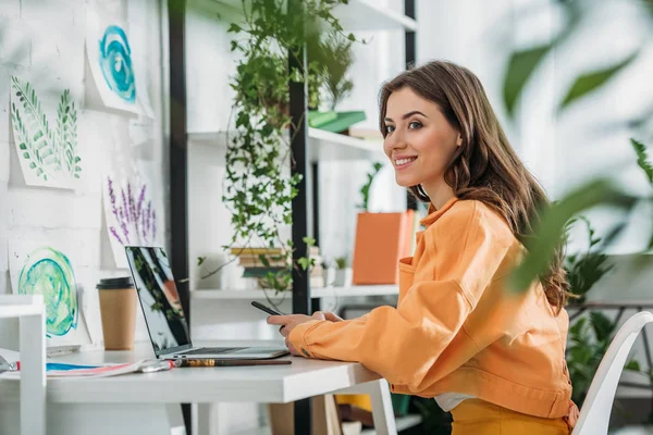Selective focus of smiling young woman using smartphone while sitting at desk near laptop — Stock Photo