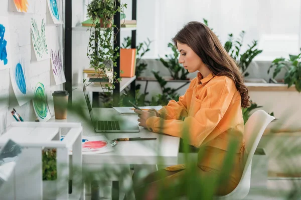 Selective focus of thoughtful young woman sitting at desk near laptop and using smartphone — Stock Photo