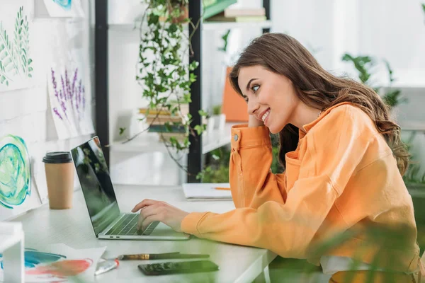 Foco seletivo de alegre jovem mulher sentada na mesa, sorrindo e usando laptop — Fotografia de Stock