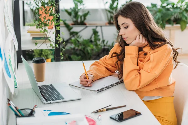 Nachdenkliche junge Frau schreibt in Notizbuch, während sie am Schreibtisch sitzt, trägt Laptop und Smartphone — Stockfoto