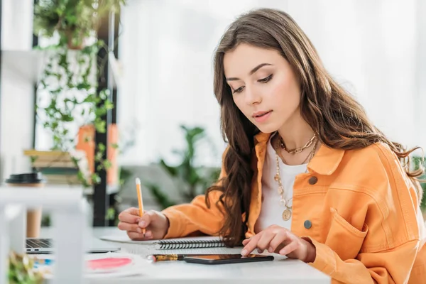 Mujer joven y atenta sentada en el escritorio, usando un teléfono inteligente y escribiendo en un cuaderno - foto de stock