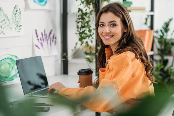 Selective focus of cheerful girl smiling at camera while using laptop and holding coffee cup — Stock Photo
