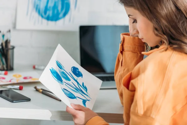 Jovem mulher pensativo sentado na mesa e olhando para a pintura com flores azuis — Fotografia de Stock