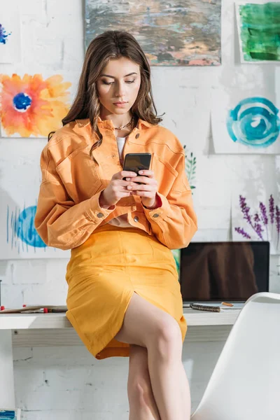 Thoughtful girl using smartphone while sitting on table near laptop with blank screen — Stock Photo