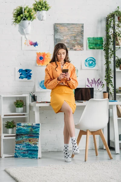 Élégante jeune femme en utilisant smartphone dans une chambre spacieuse décorée avec des plantes vertes et des peintures colorées sur le mur — Photo de stock
