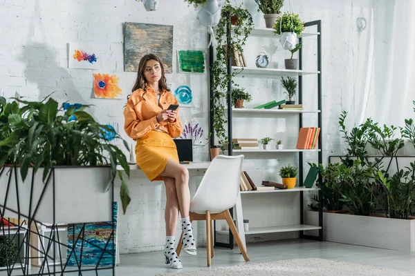 Elegante grl utilizzando smartphone in camera spaziosa decorata con piante verdi e dipinti colorati a parete — Foto stock