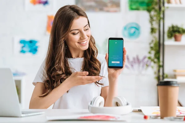 KYIV, UCRANIA - 17 DE ABRIL DE 2019: Mujer joven y atractiva sonriendo y señalando con la mano al teléfono inteligente con la aplicación de Twitter en la pantalla . - foto de stock