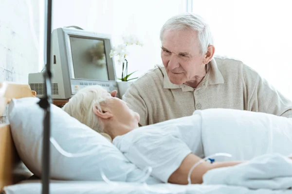 Sick senior woman with husband looking at each other in ward in clinic — Stock Photo