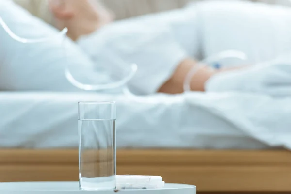 Messa a fuoco selettiva della donna anziana malata sdraiata sul letto in clinica con un bicchiere d'acqua in primo piano — Foto stock