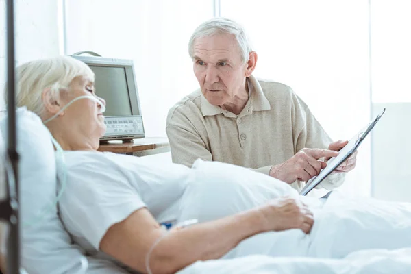 Hombre mayor sosteniendo portapapeles y mirando a la esposa enferma en la clínica - foto de stock