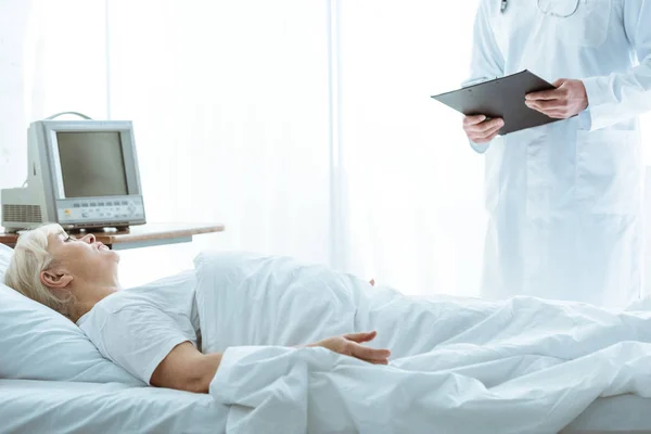 Ausgeschnittene Ansicht von Arzt mit Klemmbrett und kranker Seniorin auf Bett liegend — Stockfoto