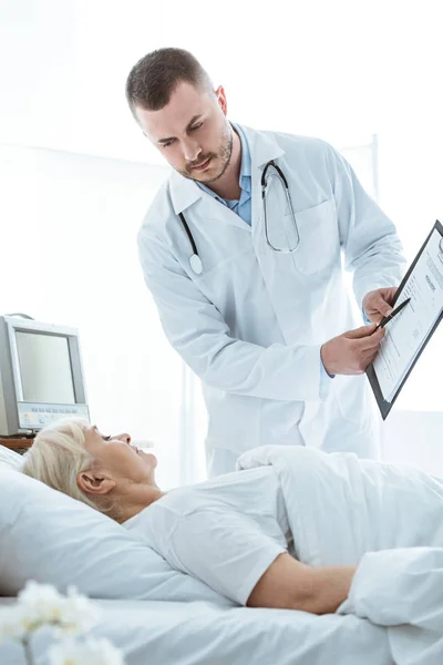 Doctor con portapapeles y anciana enferma acostada en la cama - foto de stock