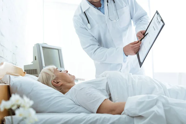 Vista ritagliata del medico con appunti e malata donna anziana sdraiata sul letto — Foto stock