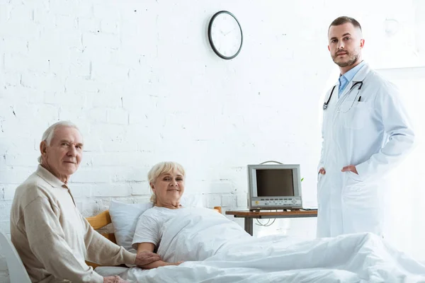 Doctor en bata blanca, hombre mayor y mujer enferma sonriente acostado en la cama en la clínica - foto de stock
