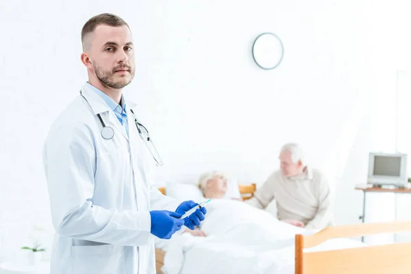 Médico en guantes de látex que sostiene la jeringa, paciente enfermo y hombre mayor en la clínica - foto de stock