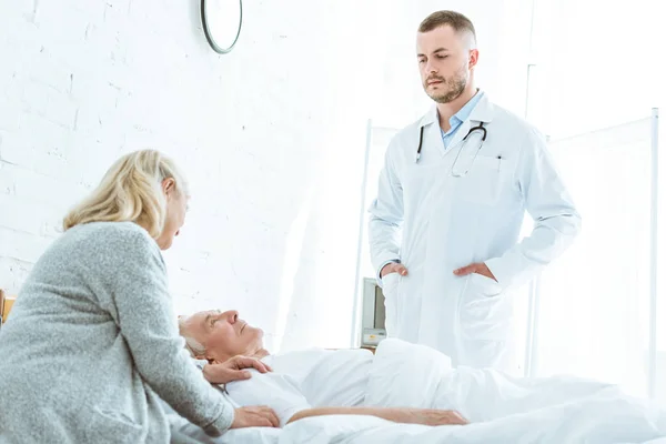 Ernsthafter Arzt steht mit Händen in den Hosentaschen neben kranker Patientin und Seniorin in Klinik — Stockfoto
