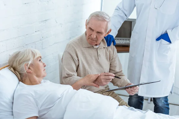 Vista parziale del medico, uomo anziano firma appunti e malato paziente sul letto — Foto stock