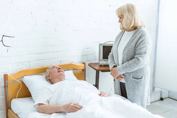 Mujer mayor de pie cerca de marido enfermo en el hospital - foto de stock