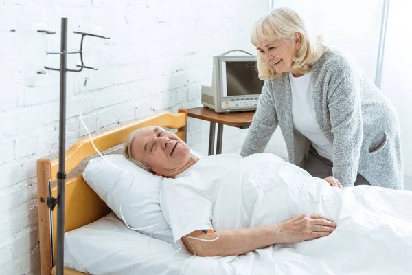 Sonriente mujer mayor de pie cerca de marido enfermo en el hospital - foto de stock