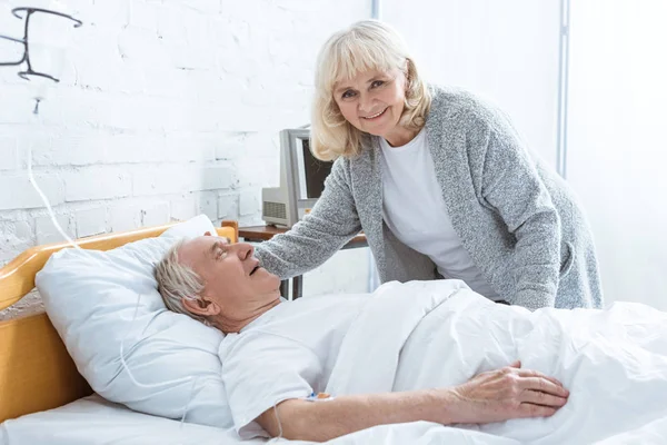 Femme âgée souriante debout près du mari malade à l'hôpital — Photo de stock