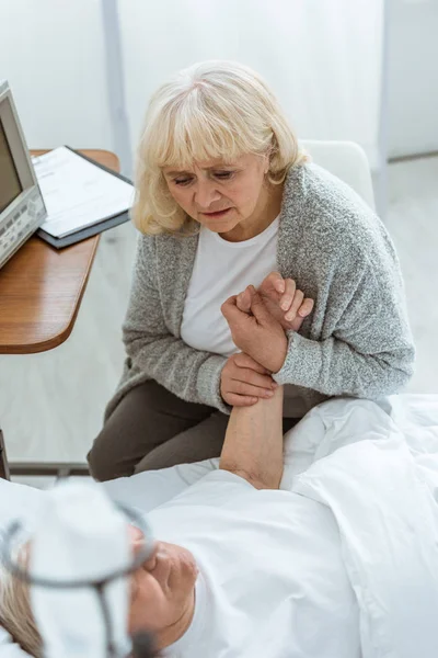 Blick auf besorgte Seniorin, die neben krankem Mann sitzt und seine Hand im Krankenhaus hält — Stockfoto