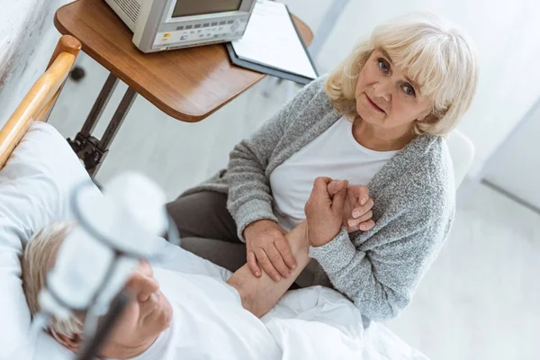 Besorgte Seniorin sitzt neben krankem Mann und hält seine Hand im Krankenhaus — Stockfoto