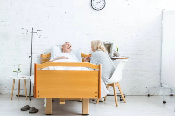 Besorgte Seniorin sitzt neben krankem Mann und hält seine Hand im Krankenhaus — Stockfoto