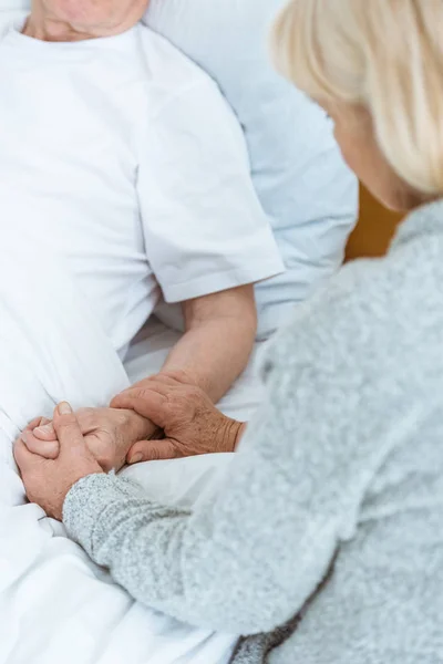 Vista recortada de la mujer mayor de la mano con el marido enfermo en el hospital - foto de stock