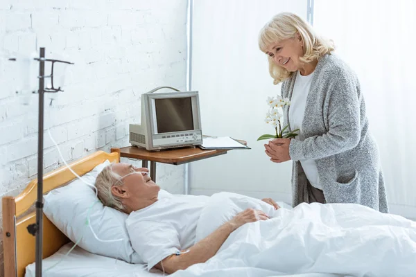 Femme âgée souriante avec des orchidées et malade à l'hôpital — Photo de stock