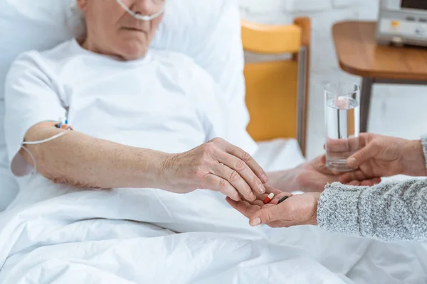 Vista recortada de la mujer mayor dando vaso de agua y pastillas a marido enfermo en el hospital — Stock Photo