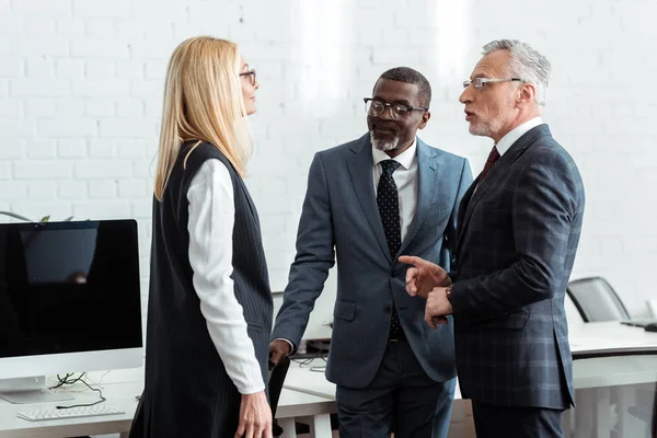Handsome businessman in glasses pointing with finger while talking with woman near african american man — Stock Photo
