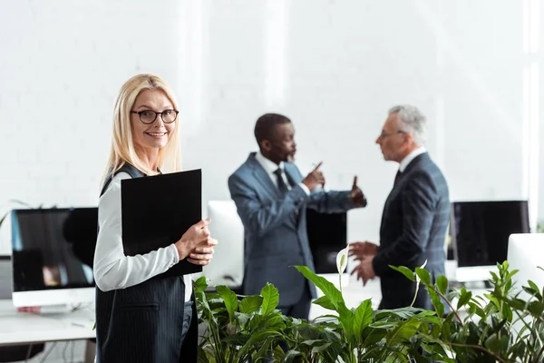 Selective focus of attractive blonde businesswoman holding clipboard while standing near multicultural businessmen — Stock Photo