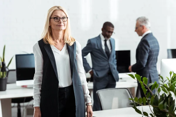 Selective focus of cheerful blonde woman standing in office near multicultural businessmen — Stock Photo