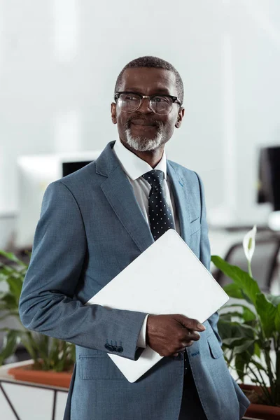 Alegre afroamericano hombre en gafas sosteniendo portátil en la oficina - foto de stock
