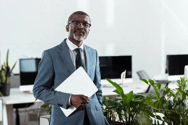 Hombre de negocios afroamericano feliz en gafas que sostienen el ordenador portátil en la oficina - foto de stock