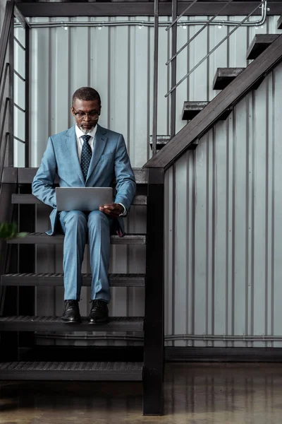 Guapo afroamericano hombre en gafas usando portátil mientras está sentado en las escaleras en la oficina - foto de stock