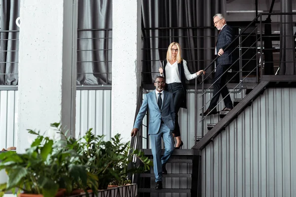Feliz rubia mujer de negocios caminando en las escaleras cerca de los hombres de negocios multiculturales - foto de stock