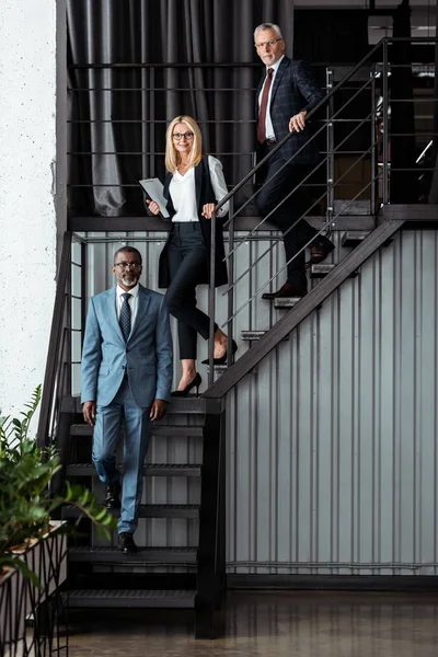 Веселая блондинка-бизнесмен, идущая по лестнице рядом с мультикультурными бизнесменами — стоковое фото