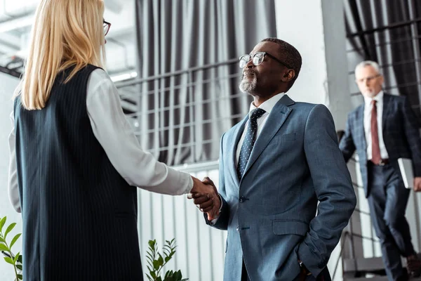 Vista de ángulo bajo de la mujer de negocios rubia estrechando la mano con hombre de negocios afroamericano - foto de stock