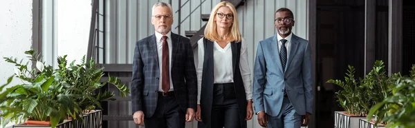 Plan panoramique de femme d'affaires blonde confiante debout avec de beaux partenaires multiculturels au bureau — Photo de stock