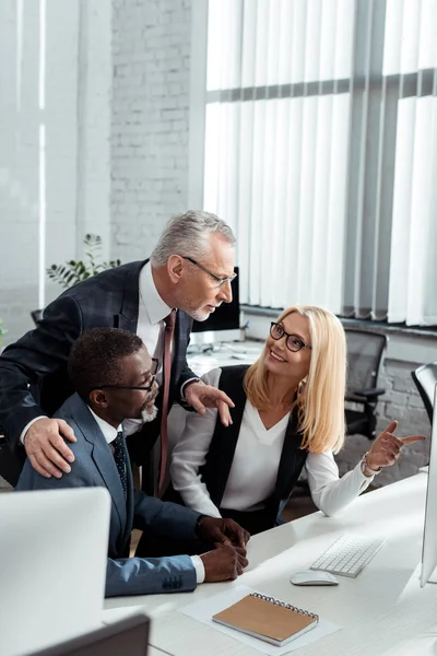 Alegre mujer de negocios mirando guapo hombre de negocios cerca de africano americano socio en la oficina - foto de stock